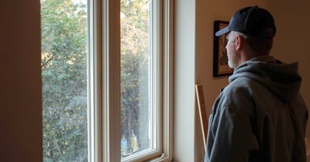 Person in a cap and hoodie standing indoors and looking out a large window.